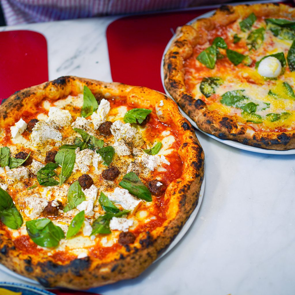Two signature pizzas from Pizzeria Carmnella: the Filetto, topped with cherry tomatoes from Mount Vesuvius, fior di latte, fresh basil, and Pecorino with no tomato sauce base, and the Elena Ferrante, layered with fior di latte, mini meatballs, ricotta, black pepper, and olive oil. A showcase of the best pizza in the world